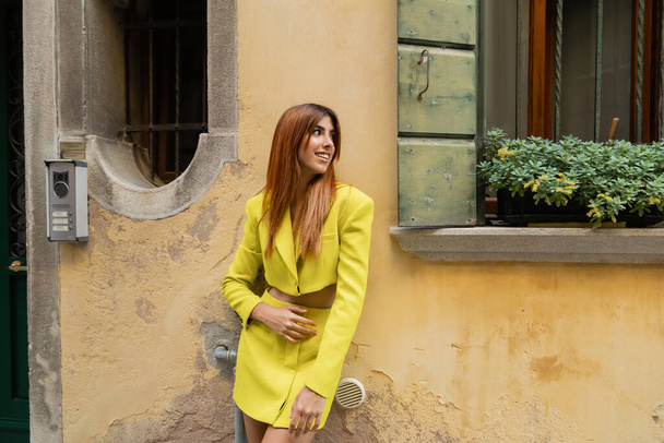 mulher alegre no terno amarelo que olha perto da janela decorada com flores potted em Veneza - Foto, Imagem