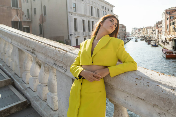молодая женщина в жёлтом костюме опирается на балюстраду моста через Большой канал в Венеции - Фото, изображение