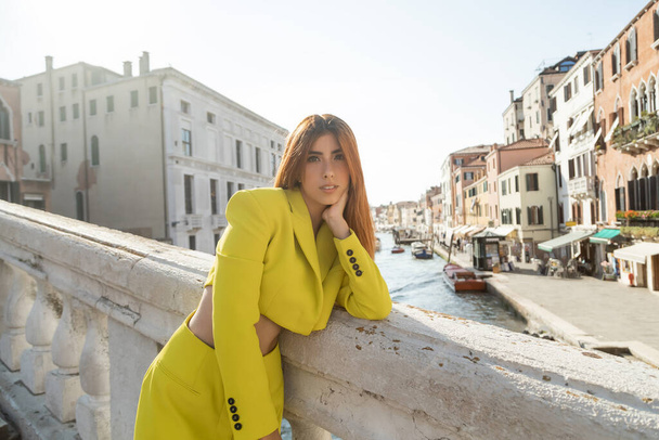 модная рыжая женщина смотрит в камеру, стоя на мосту через венецианский Гранд-канал - Фото, изображение