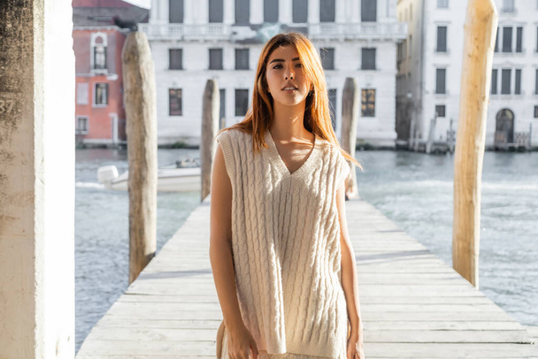 jonge vrouw in mouwloze trui op zoek naar camera in de buurt wazig pier in Venetië - Foto, afbeelding