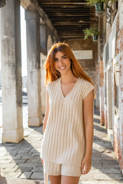 pozytywna kobieta z rudymi włosami uśmiechnięta do kamery na ulicy w Wenecji - Zdjęcie, obraz