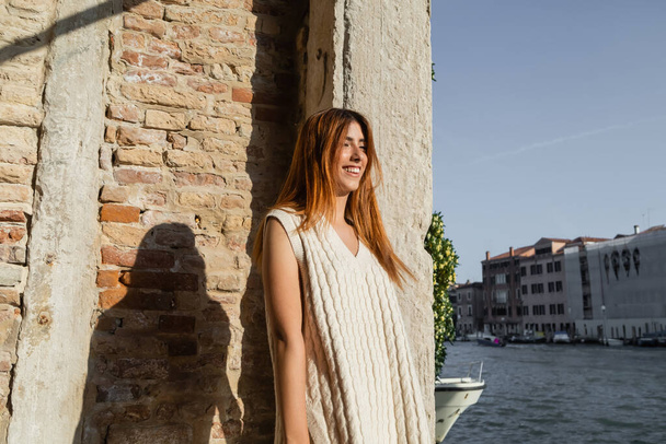 cheerful redhead woman in sleeveless jumper smiling near stone wall in Venice - Foto, imagen