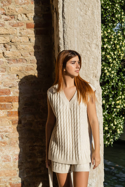 stylische Frau in ärmellosem Pullover, die in der Nähe einer Steinmauer in Venedig wegschaut - Foto, Bild
