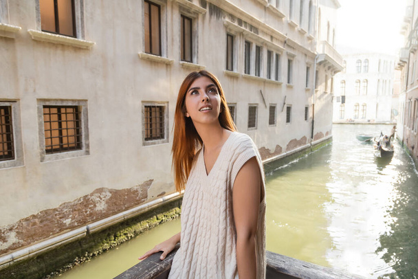 Lächelnde Frau im ärmellosen Pullover, die in der Nähe von Gebäuden und Kanälen in Venedig wegschaut - Foto, Bild
