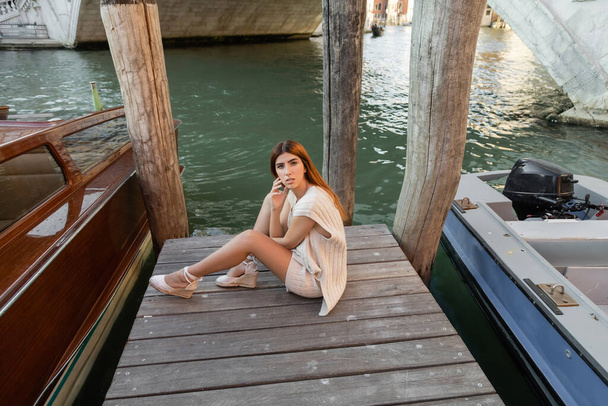 Hochwinkelaufnahme einer Frau in sommerlicher Strickbekleidung, die auf einem hölzernen Pier in Venedig sitzt - Foto, Bild