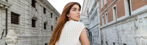 redhead woman in knitwear looking away near medieval prison in Venice, banner - Photo, Image