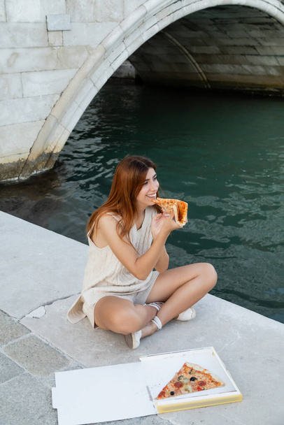Venedik 'te kanal kenarında oturmuş geleneksel pizza yiyen neşeli bir kadın. - Fotoğraf, Görsel