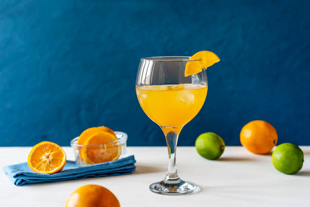 Coquetel de pássaro amarelo com rum, suco de laranja e limão em vidro com fatia de laranja, frutas, guardanapo azul na mesa branca. Fundo azul escuro. Estilo mediterrânico ou marinho. Espaço de cópia - Foto, Imagem