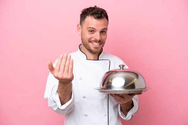 Young Brazilian chef with tray isolated on pink background inviting to come with hand. Happy that you came - Photo, image