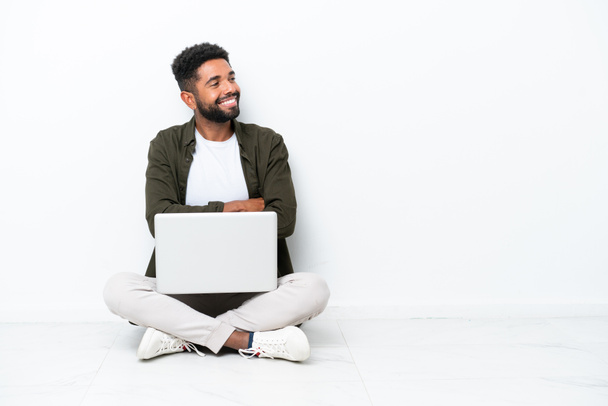 Young Brazilian man with a laptop sitting on the floor isolated on white happy and smiling - Photo, Image