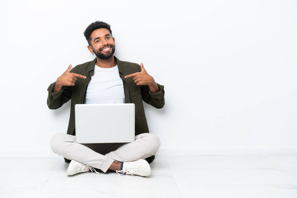 Yerde dizüstü bilgisayarı olan genç Brezilyalı bir adam kendini beğenmiş ve gururlu bir beyazın üzerinde izole bir şekilde oturuyor. - Fotoğraf, Görsel