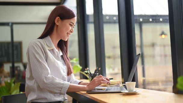 Вид збоку весела азіатська жінка сидить в сучасному кафе і працює онлайн з комп'ютерним ноутбуком
. - Фото, зображення