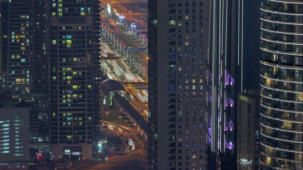 Highway and overpass between skyscrapers with glowing windows in Dubai downtown aerial night timelapse. Glowing windows in towers near metro station and busy traffic from above. - Fotoğraf, Görsel