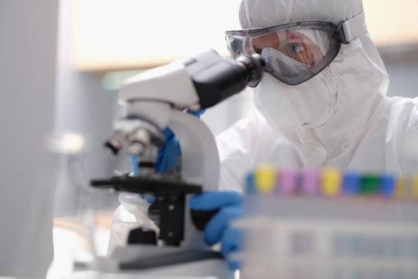 Ученый в защитном костюме, смотрящий через микроскоп в современной оборудованной лаборатории. Лаборант, изучающий эволюцию вируса с использованием высоких технологий для научных исследований - Фото, изображение