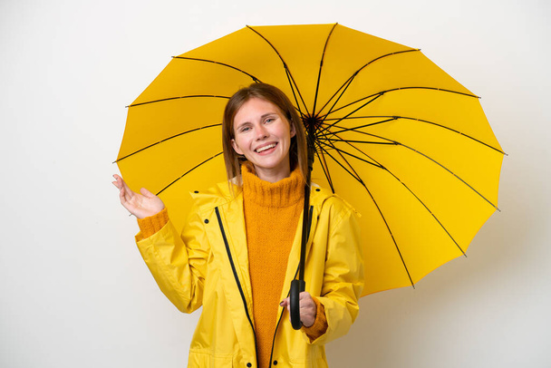Mujer inglesa joven con abrigo impermeable y paraguas aislados sobre fondo blanco extendiendo las manos a un lado para invitar a venir - Foto, imagen
