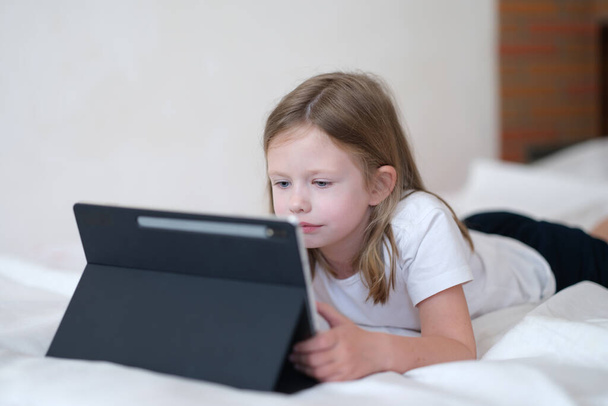 Klein meisje kijkt zorgvuldig naar de tablet terwijl ze op bed ligt. Mobiel leren apps voor kinderen concept - Foto, afbeelding
