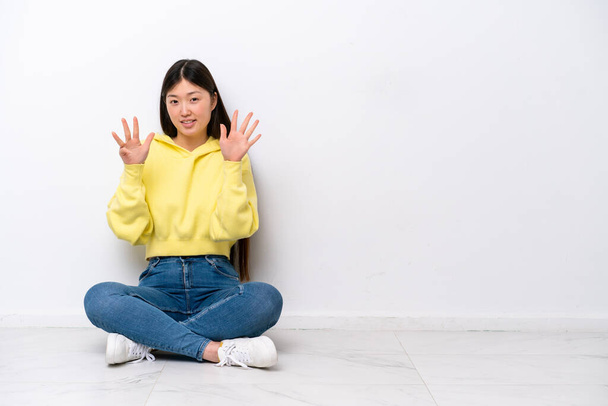 Молодая китаянка сидит на полу изолированный на белой стене, считая девять с пальцами - Фото, изображение