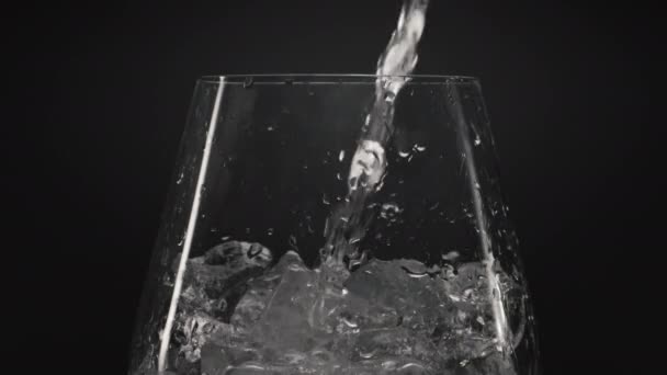 IJswater gieten wijnglas close-up. Kristalheldere bellenvloeistof met ijsblokjes in transparant glas. Minerale aqua met lucht druppels zwarte achtergrond. Verfrissend cocktailconcept voorbereiden - Video