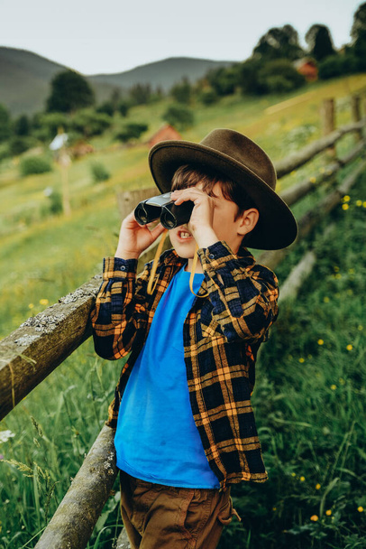 Мальчик смотрит сквозь бинокль, стоя летом на вершине холма. Семейное путешествие в горы летом. Мальчик рассматривает горный пейзаж через бинокль летом. - Фото, изображение
