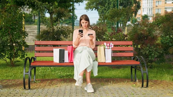 Кавказька молода жінка сидить на лавці в парку з сумками для покупок і тримає кредитну картку та телефон. - Фото, зображення