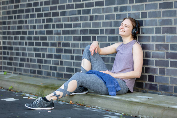 A young Caucasian woman with earphones takes a break after her fitness workout leaning on a stone wall - Foto, Bild