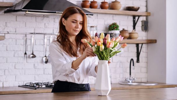 Valkoihoinen nainen katselee värikkäitä tulppaaneja maljakossa kodikas minimalistinen keittiö ympäristössä - Valokuva, kuva