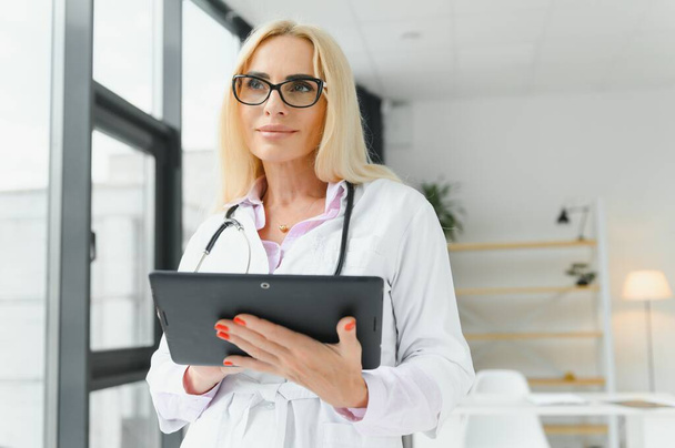 Portrait of beautiful mature woman doctor holding digital tablet. Confident female doctor using digital tablet with colleague talking in background at hospital - Photo, Image