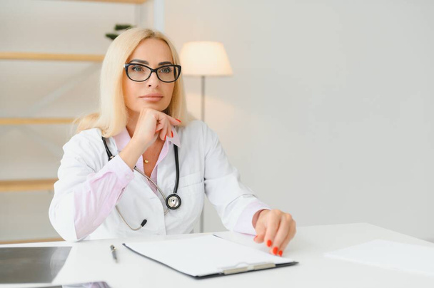 Portret lekarza w średnim wieku nosi biały płaszcz lekarski ze stetoskopem na szyi. Uśmiechnięty lekarz stojący w prywatnej klinice. - Zdjęcie, obraz