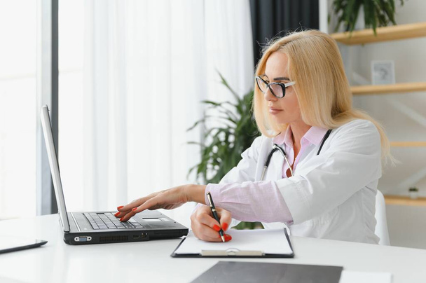 Portraitaufnahme einer Ärztin mittleren Alters, die am Schreibtisch sitzt und in der Arztpraxis arbeitet. - Foto, Bild