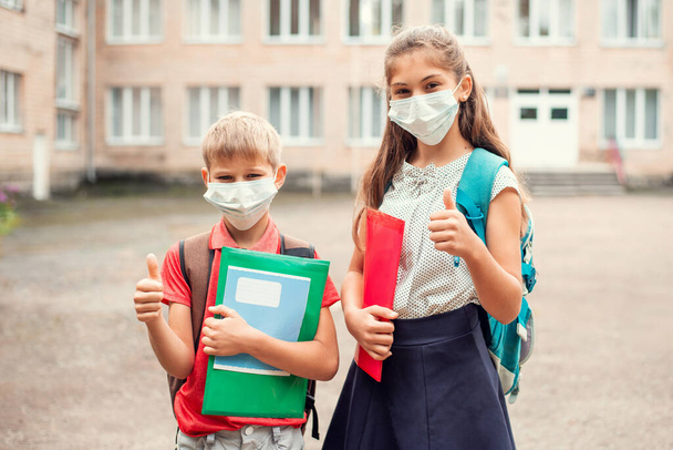 Bambini in maschera medica prima delle lezioni mostrando i pollici in alto, in piedi con gli zaini pronti per lo studio - Foto, immagini