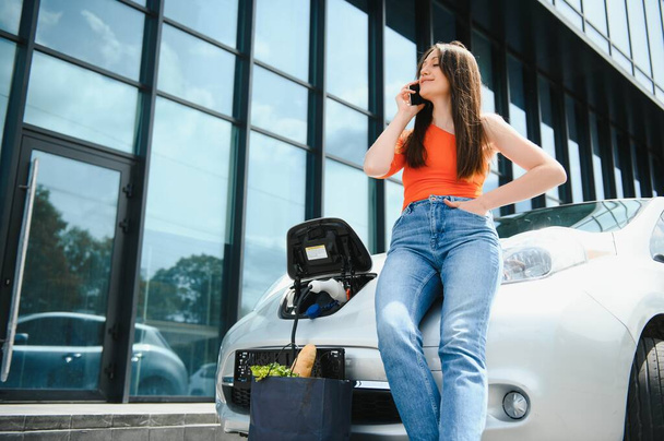 Young woman is standing near the electric car and looks at the smart phone. The rental car is charging at the charging station for electric vehicles. Car sharing - Foto, immagini
