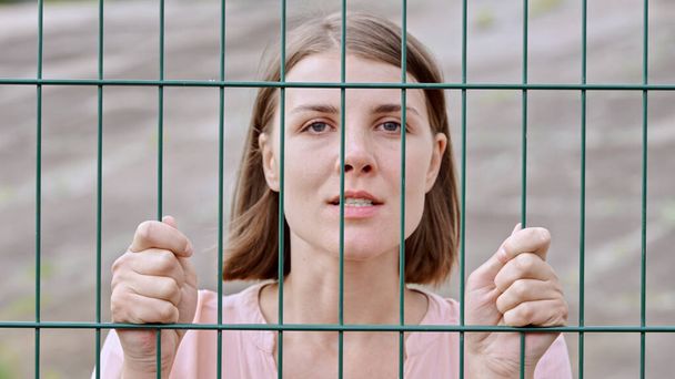 Caucasian young woman looking at the camera and standing behind a fence with her hands on the bars - Foto, immagini