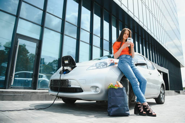 Usar el teléfono inteligente mientras espera. Mujer en la estación de carga de coches eléctricos durante el día. Vehículo nuevo - Foto, Imagen