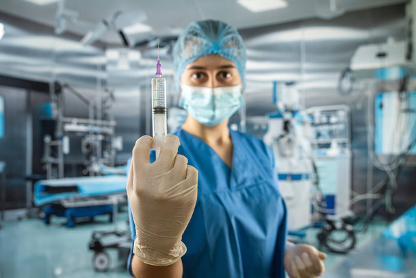 女性医師やアシスタントは、手術室で薬で注射器を保持ウイルスからの完全な保護のための手袋、フェイスマスク眼鏡や帽子と青の制服を着用  - 写真・画像