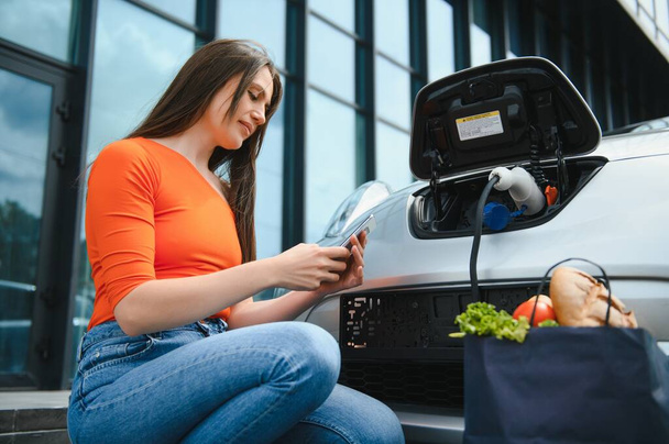Genç kadın elektrikli arabanın yanında duruyor ve akıllı telefona bakıyor. Kiralık araba elektrikli araçlar için şarj istasyonunda şarj oluyor. Araba paylaşımı - Fotoğraf, Görsel