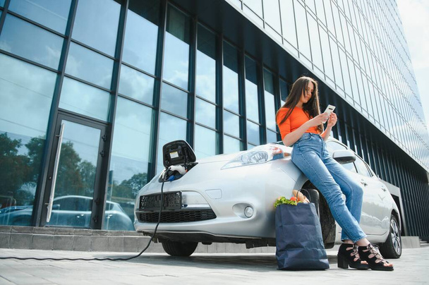 Young woman is standing near the electric car and looks at the smart phone. The rental car is charging at the charging station for electric vehicles. Car sharing - Fotografie, Obrázek
