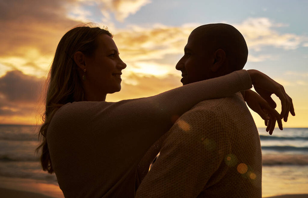 Loving young affectionate interracial couple in silhouette facing each other while spending time together on the beach at sunset. Lovers sharing romantic moment against golden sky while on vacation. - Photo, Image