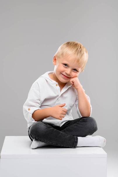 A small fair-haired boy in a shirt and pants sits on a white cube. Portrait. Spectrum of emotions - 写真・画像