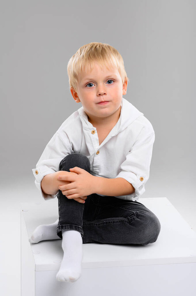 A small fair-haired boy in a shirt and pants sits on a white cube. Portrait. Spectrum of emotions - Foto, Bild