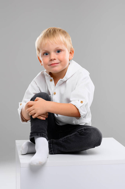 Un pequeño chico de pelo rubio con camisa y pantalones se sienta en un cubo blanco. Retrato. Espectro de emociones - Foto, Imagen
