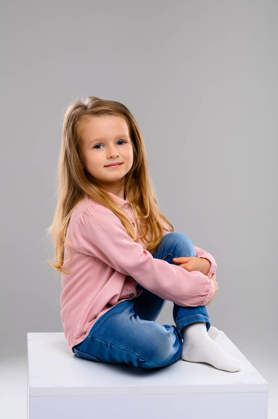 Cute blonde girl in a pink sweater and jeans on a white cube in the studio. Portrait. beautiful girl - Fotografie, Obrázek