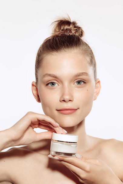 Half-length portrait of charming young girl without makeup holding cream bottle isolated over white studio background. Natural cosmetics treatment. Concept of beauty, skincare, cosmetology, spa - Photo, image