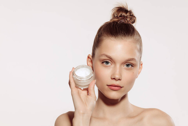 Half-length portrait of charming young girl without makeup holding cream bottle isolated over white studio background. Natural cosmetics treatment. Concept of beauty, skincare, cosmetology, spa - Φωτογραφία, εικόνα