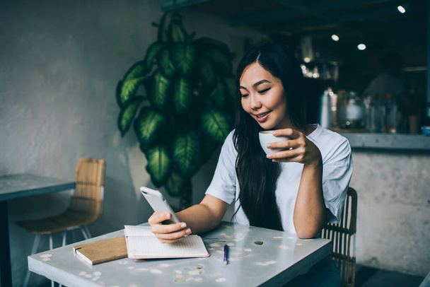 Delighted Asian female drinking hot beverage and text messaging on cellphone while sitting at at with notebooks in modern cafe - Фото, изображение
