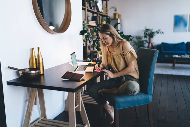 European girl writing something in adhesive paper with pen at home. Concept of remote or e-learning. Young focused woman sitting in armchair at desk with opened laptop. Interior of studio apartment - 写真・画像