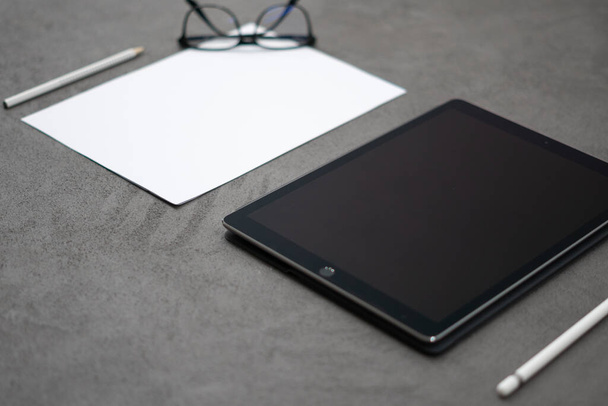 A closeup of a black thin tablet with a white pen and eyeglasses on the gray fabric - Foto, afbeelding