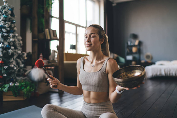European girl meditating with standing bell on fitness mat at home. Concept of healthy lifestyle. Young thoughtful pretty athletic woman with closed eyes in sportswear. Interior of studio apartment - Фото, изображение