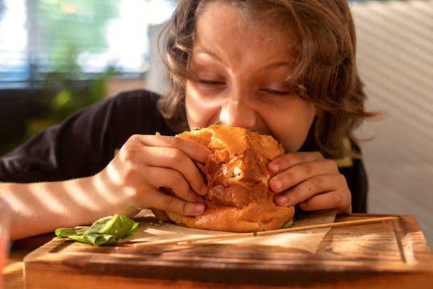  chico muerde hamburguesa caliente con chuleta de cerdo, lechuga, tomate, pepinillo en el interior. Día de verano - Foto, imagen