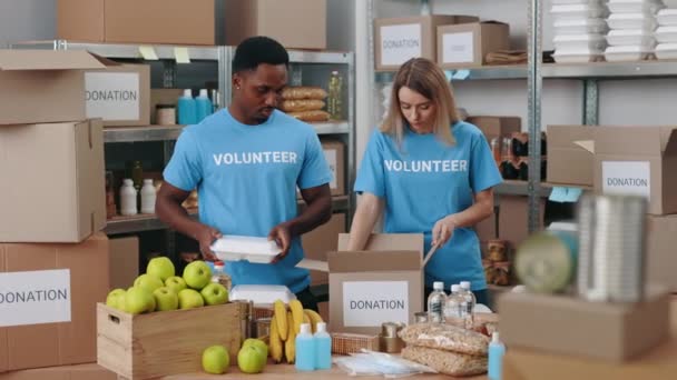 Beyaz kadın ve Afro-Amerikan erkek depoda birlikte duruyor ve bağış için kutuları paketliyorlar. Gıda bankasında gönüllü çalışan çok ırklı insanlar.. - Video, Çekim