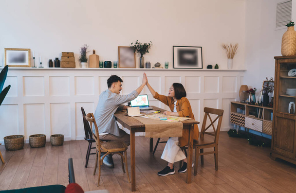 Aasialainen nainen ja valkoihoinen mies antavat viisi toisiaan pöydässä kotona. Nuori monirotuinen pari suunnittelee tulevaa viikonloppua, lomaa tai matkaa. Puinen pöytä kartalla, digitaaliset laitteet ja eri kohteita - Valokuva, kuva
